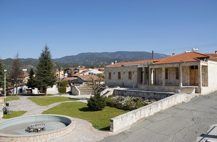 View of Trimiklini village square with traditional stone buildings and Troodos foothills in the background